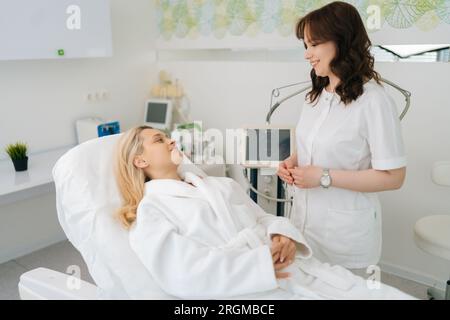 Portrait de cosmétologue souriante parlant avec la jeune femme cliente en peignoir blanc couché sur le canapé avant la procédure rf-lifting dans la beauté moderne Banque D'Images