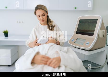 Portrait de cosmétologue féminin concentré faisant massage de levage de visage d'échographie avec l'équipement professionnel pour jeune femme couchée sur le canapé Banque D'Images