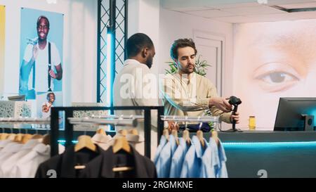 Homme afro-américain payant pour de nouveaux vêtements modernes, faisant la transaction sur le terminal de pos dans le centre commercial. Client masculin parlant à l'assistant de magasin à la caisse enregistreuse, activité commerciale. Prise de vue à main levée. Banque D'Images