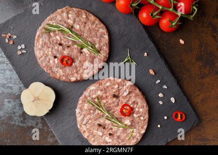 Viandes de hamburger de bœuf haché cru, romarin, légumes et épices sur une assiette de pierre noire sur un fond sombre, vue de dessus. Viande semi-finie Banque D'Images