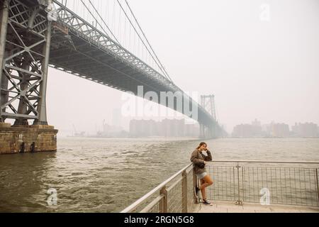 Adolescente posant sous le pont Williamsburg dans Domino Park, Brooklyn, dans une journée brumeuse et brumeuse. Paysage grand angle. Banque D'Images