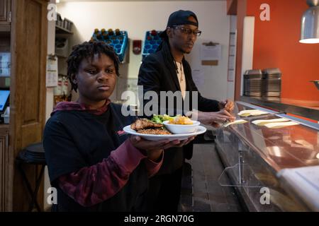 Reportage : SNAP emploi et formation au café Reconcile à la Nouvelle-Orléans LA (février 2023) - Right, un formateur principal défenseur au café Reconcile, un restaurant soul food à la Nouvelle-Orléans, aide à servir de la nourriture et conseille un stagiaire, 16 février 2023. Banque D'Images