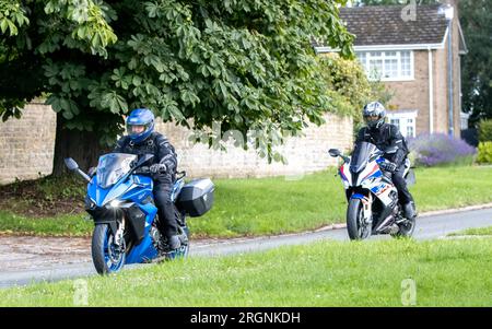 Whittlebury, Northants., Royaume-Uni - 6 août 2023. Moto Suzuki bleue suivie d'une moto BMW blanche traversant un village anglais un dimanche Banque D'Images