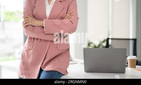 Image recadrée d'une femme d'affaires confiante dans un costume d'affaires rose assis sur une table avec ses bras croisés Banque D'Images