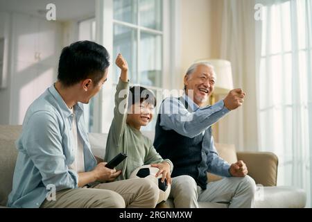 Fils asiatique père grand-père assis sur le canapé à la maison célébrant le but et la victoire tout en regardant la diffusion en direct du match de football à la télévision ensemble Banque D'Images