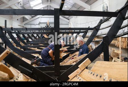 David Steptoe et Mike Pratt travaillent sur la réplique du Sutton Hoo, au Longshed à Woodbridge, Suffolk. Trois apprentis menuisiers entreprennent un stage de deux semaines avec la Sutton Hoo Ship's Company, qui dirige les travaux sur la réplique grandeur nature de 88 pieds de long. Date de la photo : Vendredi 11 août 2023. Banque D'Images