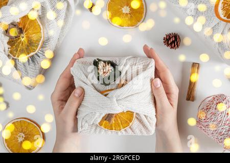 Boîte cadeau en tissu blanc. Femmes emballage des mains cadeau durable sur table blanche avec éléments décoratifs. Orange séché, cannelle, Cones. Pas de déchets CH Banque D'Images