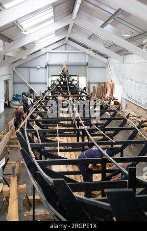 Les volontaires travaillent sur la réplique du navire Sutton Hoo, au Longshed à Woodbridge, dans le Suffolk. Trois apprentis menuisiers entreprennent un stage de deux semaines avec la Sutton Hoo Ship's Company, qui dirige les travaux sur la réplique grandeur nature de 88 pieds de long. Date de la photo : Vendredi 11 août 2023. Banque D'Images