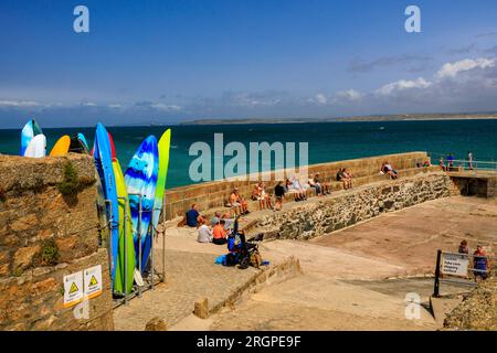 Planches de surf colorées et touristes à l'abri du vent sur un brise-lames à St Ives, Cornouailles, Angleterre, Royaume-Uni Banque D'Images