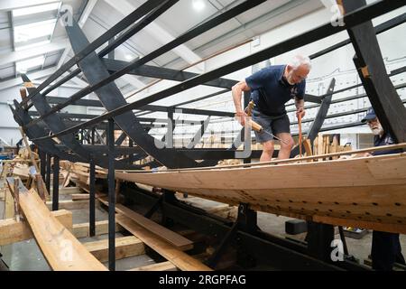 Mike Pratt et David Steptoe travaillent sur la réplique du Sutton Hoo, au Longshed à Woodbridge, Suffolk. Trois apprentis menuisiers entreprennent un stage de deux semaines avec la Sutton Hoo Ship's Company, qui dirige les travaux sur la réplique grandeur nature de 88 pieds de long. Date de la photo : Vendredi 11 août 2023. Banque D'Images