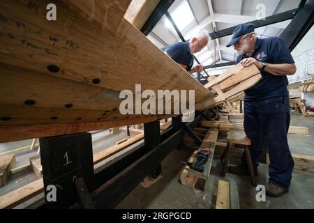 Mike Pratt et David Steptoe travaillent sur la réplique du Sutton Hoo, au Longshed à Woodbridge, Suffolk. Trois apprentis menuisiers entreprennent un stage de deux semaines avec la Sutton Hoo Ship's Company, qui dirige les travaux sur la réplique grandeur nature de 88 pieds de long. Date de la photo : Vendredi 11 août 2023. Banque D'Images