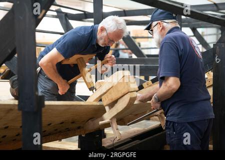 Mike Pratt et David Steptoe travaillent sur la réplique du Sutton Hoo, au Longshed à Woodbridge, Suffolk. Trois apprentis menuisiers entreprennent un stage de deux semaines avec la Sutton Hoo Ship's Company, qui dirige les travaux sur la réplique grandeur nature de 88 pieds de long. Date de la photo : Vendredi 11 août 2023. Banque D'Images