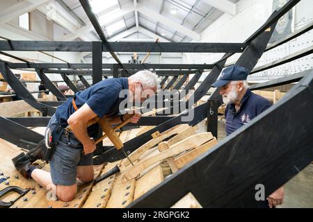 Mike Pratt et David Steptoe travaillent sur la réplique du Sutton Hoo, au Longshed à Woodbridge, Suffolk. Trois apprentis menuisiers entreprennent un stage de deux semaines avec la Sutton Hoo Ship's Company, qui dirige les travaux sur la réplique grandeur nature de 88 pieds de long. Date de la photo : Vendredi 11 août 2023. Banque D'Images