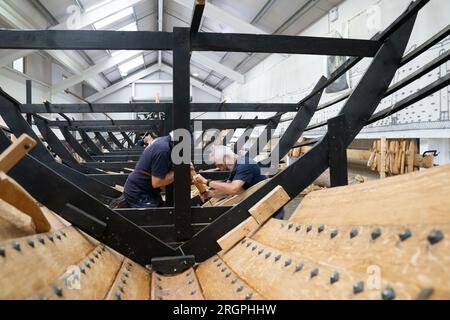 David Steptoe et Mike Pratt travaillent sur la réplique du Sutton Hoo, au Longshed à Woodbridge, Suffolk. Trois apprentis menuisiers entreprennent un stage de deux semaines avec la Sutton Hoo Ship's Company, qui dirige les travaux sur la réplique grandeur nature de 88 pieds de long. Date de la photo : Vendredi 11 août 2023. Banque D'Images