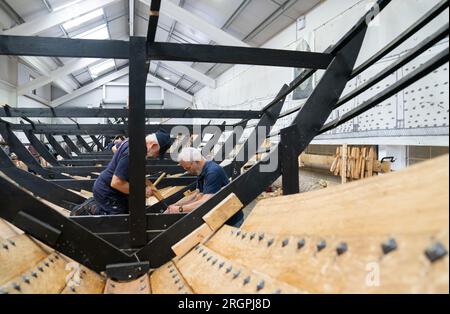 David Steptoe et Mike Pratt travaillent sur la réplique du Sutton Hoo, au Longshed à Woodbridge, Suffolk. Trois apprentis menuisiers entreprennent un stage de deux semaines avec la Sutton Hoo Ship's Company, qui dirige les travaux sur la réplique grandeur nature de 88 pieds de long. Date de la photo : Vendredi 11 août 2023. Banque D'Images