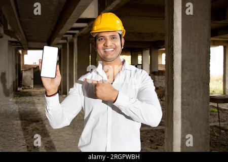 Portrait d'heureux jeune homme indien beau ingénieur civil ou architecte portant une chemise blanche et un casque de sécurité jaune tenant un téléphone intelligent avec un écran blanc Banque D'Images