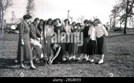 Années 1950, historique, à l'extérieur dans une zone d'herbe d'une banlieue verdoyante de l'Amérique, un nouvel arbre est planté dans une communauté, les jeunes se rassemblent pour regarder l'un de leurs propres planter un nouveau jeune arbre, USA. Banque D'Images