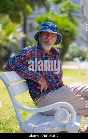 Un vieil homme avec une barbe dans un chapeau panama et une chemise à carreaux est assis sur un banc de parc par une journée ensoleillée d'été. Banque D'Images