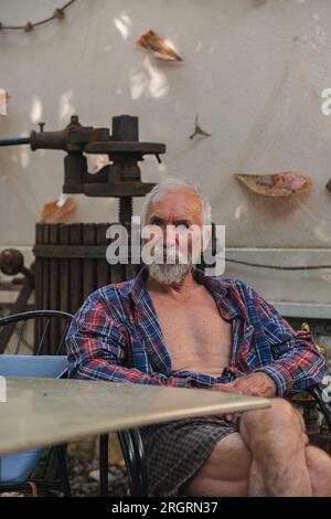 Un vieil homme dans une chemise à carreaux à la maison est assis à une table entourée par la pêche et les attributs de vin. Un homme âgé fermier et pêcheur repose dans sa propre villa. Banque D'Images