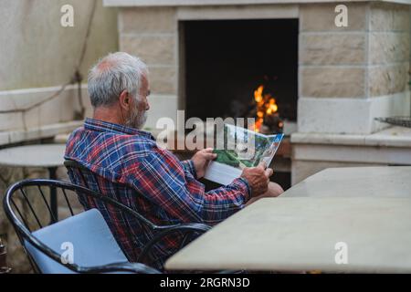 Un vieil homme dans une chemise à carreaux à la maison est assis à une table et lit le journal. Un homme âgé fermier et pêcheur repose dans sa propre villa. Banque D'Images