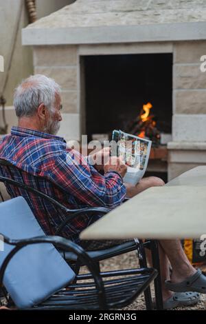 Un vieil homme dans une chemise à carreaux à la maison est assis à une table et lit le journal. Un homme âgé fermier et pêcheur repose dans sa propre villa. Banque D'Images