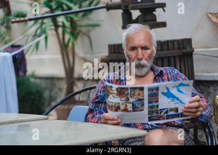 Un vieil homme dans une chemise à carreaux à la maison est assis à une table et lit le journal. Un homme âgé fermier et pêcheur repose dans sa propre villa. Banque D'Images