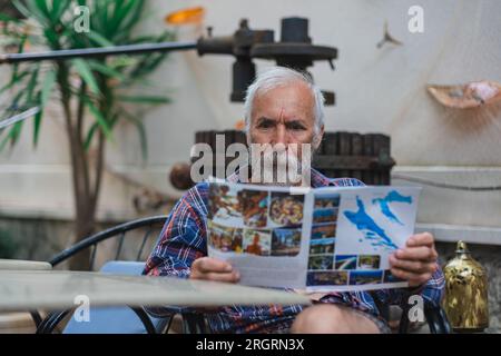 Un vieil homme dans une chemise à carreaux à la maison est assis à une table et lit le journal. Un homme âgé fermier et pêcheur repose dans sa propre villa. Banque D'Images