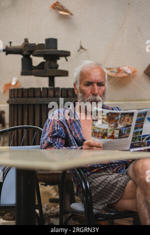 Un vieil homme dans une chemise à carreaux à la maison est assis à une table et lit le journal. Un homme âgé fermier et pêcheur repose dans sa propre villa. Banque D'Images