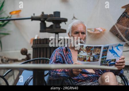 Un vieil homme dans une chemise à carreaux à la maison est assis à une table et lit le journal. Un homme âgé fermier et pêcheur repose dans sa propre villa. Banque D'Images