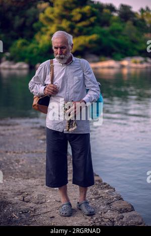 Un vieil homme avec une barbe se tient sur le bord de la mer. Un homme plus âgé voyage. Un vieil homme marche le long du bord de mer. Banque D'Images