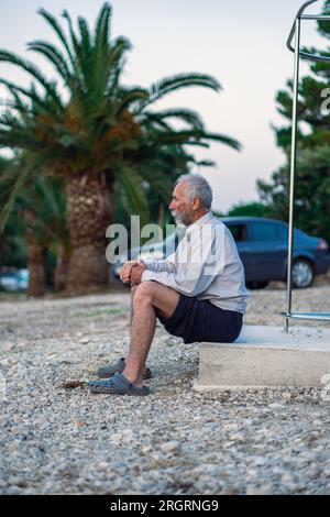 Un vieil homme sur la plage dans une chemise blanche et un pantalon foncé. Un homme âgé avec une barbe marche seul sur la plage au coucher du soleil. Portrait d'un poil gris solitaire Banque D'Images