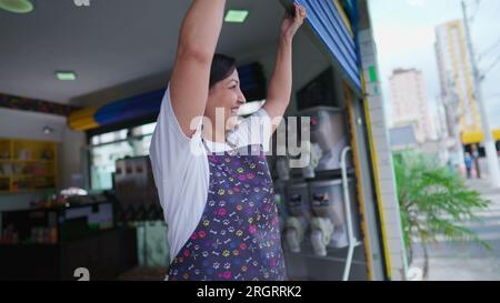 Femme propriétaire d'une vitrine de garage d'ouverture de magasin local. Jeune femme entrepreneur portant un tablier commençant la routine de la journée Banque D'Images