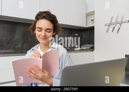 Portrait de femme brune fait ses devoirs, travaille à la maison, se prépare à l'examen dans la cuisine, s'assoit avec ordinateur portable et ordinateur portable Banque D'Images