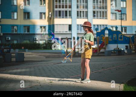 Mignonne fille d'âge scolaire souffle de grosses bulles de savon dans la cour d'un bâtiment de plusieurs étages en été. concept d'été, jeux de plein air Banque D'Images