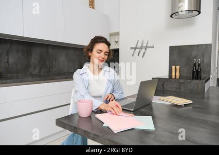 Portrait de femme brune fait ses devoirs, travaille à la maison, se prépare à l'examen dans la cuisine, s'assoit avec ordinateur portable et ordinateur portable Banque D'Images