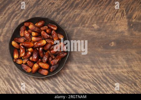 Dattes séchées dans une assiette noire sur un fond brun en bois. Fruits secs avec espace de copie. Vue de dessus, pose à plat. Banque D'Images