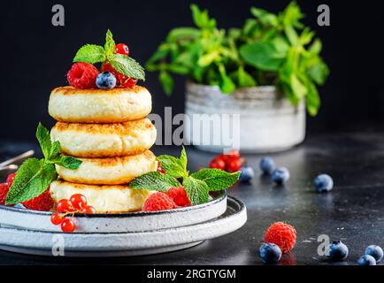 Crêpes épaisses au fromage cottage aux myrtilles, framboises et groseilles rouges, décorées de menthe fraîche sur une assiette grise, délicieux petit déjeuner. Tabl. Noir Banque D'Images