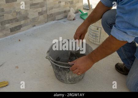 Image des mains d'un maçon mélangeant du ciment et de la colle dans un seau. Travaux manuels de maçonnerie et de carrelage. Banque D'Images