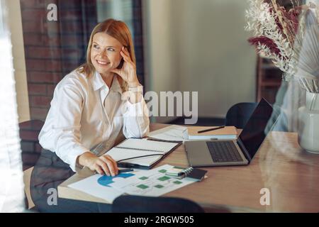 Femme analyste travaillant avec des documents et utiliser un ordinateur portable tout en étant assis dans un coworking confortable près de la fenêtre Banque D'Images