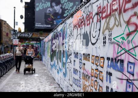 On voit des gens marcher près d'un mur avec des slogans anti-chinois dans Brick Lane. Le 5 août, les étudiants du Royal College of Arts (RCA) ont peint le mur en blanc et recouvert tous les graffitis existants des 12 valeurs fondamentales du socialisme pendant la Révolution culturelle, déclenchant un débat animé entre les artistes locaux et les communautés chinoises et remettant en question la propagation de la propagande pro-chinoise. Depuis le 8 août, le Conseil de Tower Hamlets a publié une déclaration exhortant les gens à cesser de peindre de nouveaux graffitis illégaux sur le mur, encore plus de militants anti-PCC sont arrivés et ont peint sur le mur A. Banque D'Images