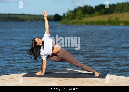 Femme pratiquant le yoga faisant l'exercice Utthita Parshvakonasana, pose d'angle latéral étendue, s'exerçant en vêtements de sport sur le bord du lac sur un morni chaud ensoleillé Banque D'Images