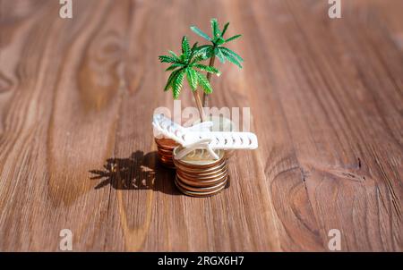 Piles de pièces de monnaie et un transat de plage jouet sous des palmiers miniatures placés sur un fond en bois sombre. Centres financiers offshore et paradis fiscal concep Banque D'Images