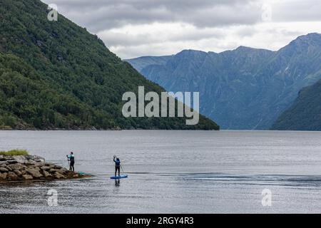 Stand up paddle femelle en combinaison de plongée pagayant sur un grand lac avec des montagnes vertes et des nuages blancs bas sur un jour de pluie brumeux à Hellesylt Norvège Banque D'Images