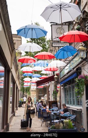 Une femme d'affaires tirant un sac à roulettes et marchant sous des parapluies colorés rouges, blancs et bleus sur Church court, Richmond, TW9, Surrey, Angleterre, ROYAUME-UNI Banque D'Images