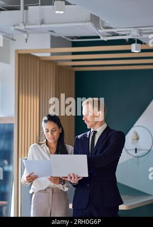 Deux employés divers regardant un ordinateur portable discuter de documents financiers au bureau. Banque D'Images