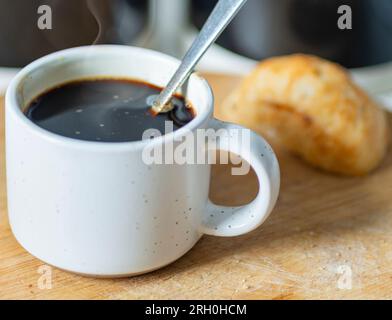 Fraîchement versé, chaud, prêt à boire, dans une tasse à café expresso et cuillère en argent utilisé pour remuer, sur une planche à découper en bois à côté d'un petit pain, sucre et co Banque D'Images