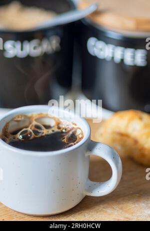 Fraîchement versé et chaud à la vapeur, prêt à boire, tasse à café sur une planche à découper en bois à côté d'un petit pain, sucre demerara et récipients à café, récents Banque D'Images