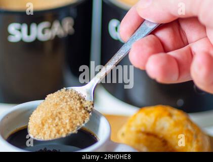 Expresso fraîchement versé, chaud et prêt à boire au petit déjeuner, une cuillère en argent sur le point de mélanger dans la cassonade pour sucrer, sur une planche de bois, un petit pain, du sucre Banque D'Images