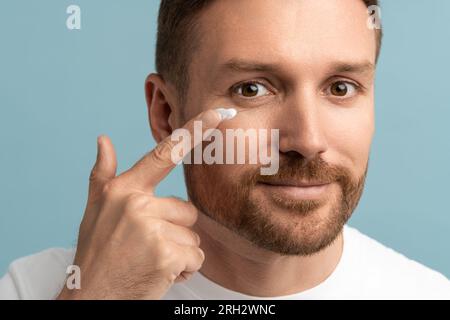 Gros plan du beau barbu homme appliquant une crème hydratante sur la peau du visage regardant la caméra. Bien-être Banque D'Images
