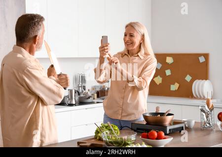 Femme mûre prenant en photo son mari avec des couteaux dans la cuisine Banque D'Images